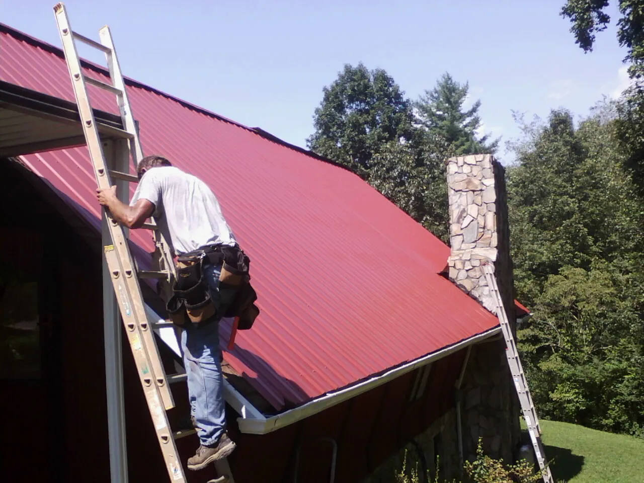 Roofer on a ladder beside a red metal roof and stone chimney — Soffit & Fascia Repair by Roofing Brothers in Bowling Green